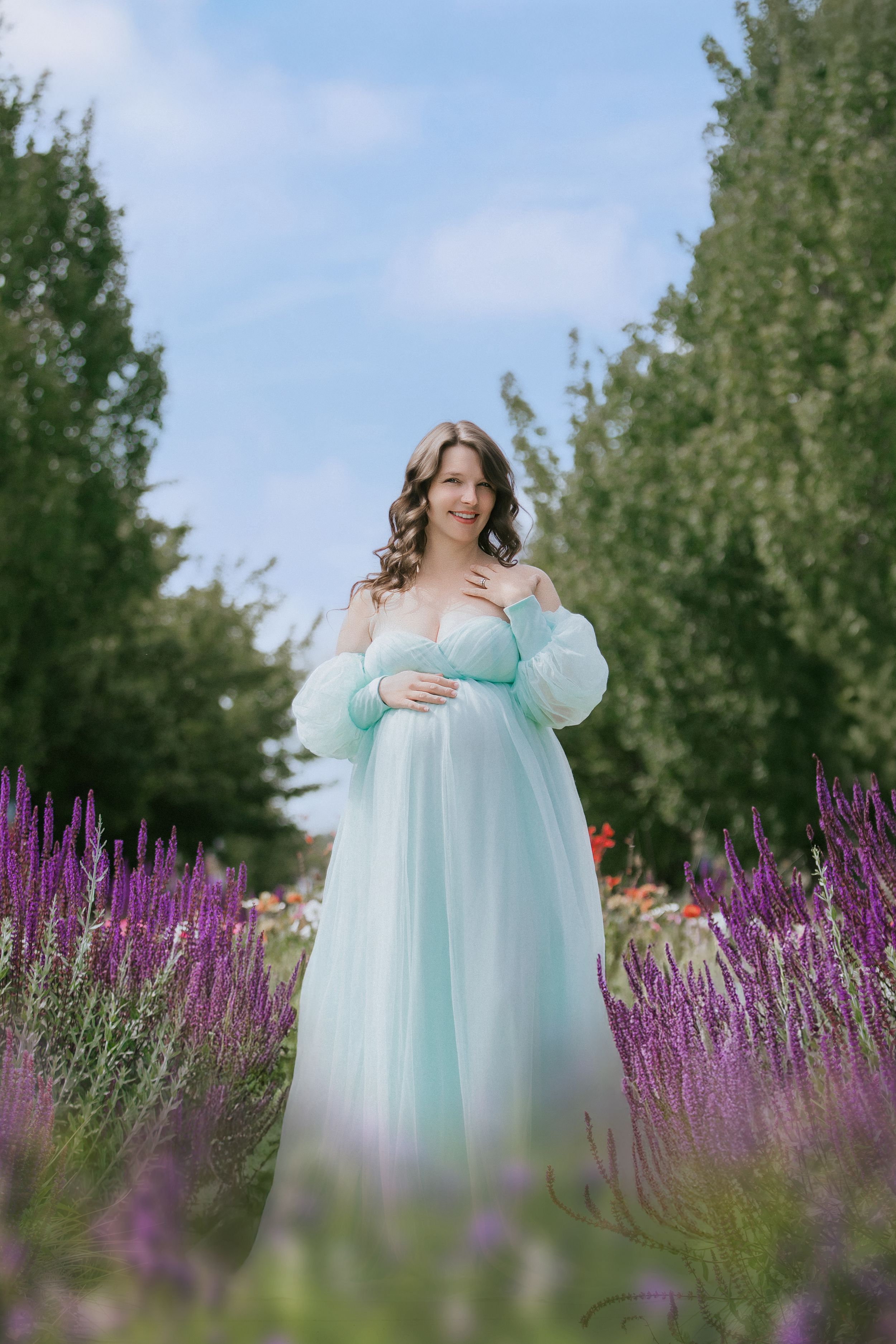 Figure in a flowing mint green dress poses gracefully among vibrant purple lavender flowers in a garden setting.