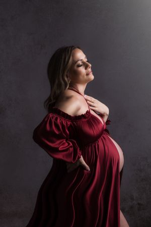 An elegant maternity portrait features a woman in a flowing burgundy off-shoulder dress against a dark background.
