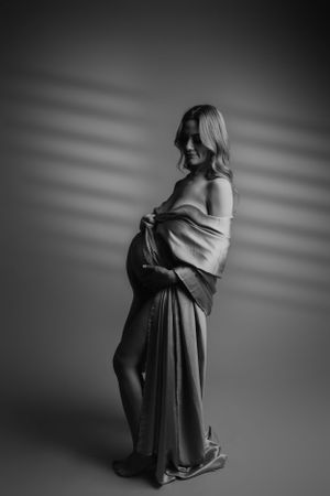 Artistic black and white maternity portrait with dramatic shadows showing an expectant mother wrapped in flowing fabric.