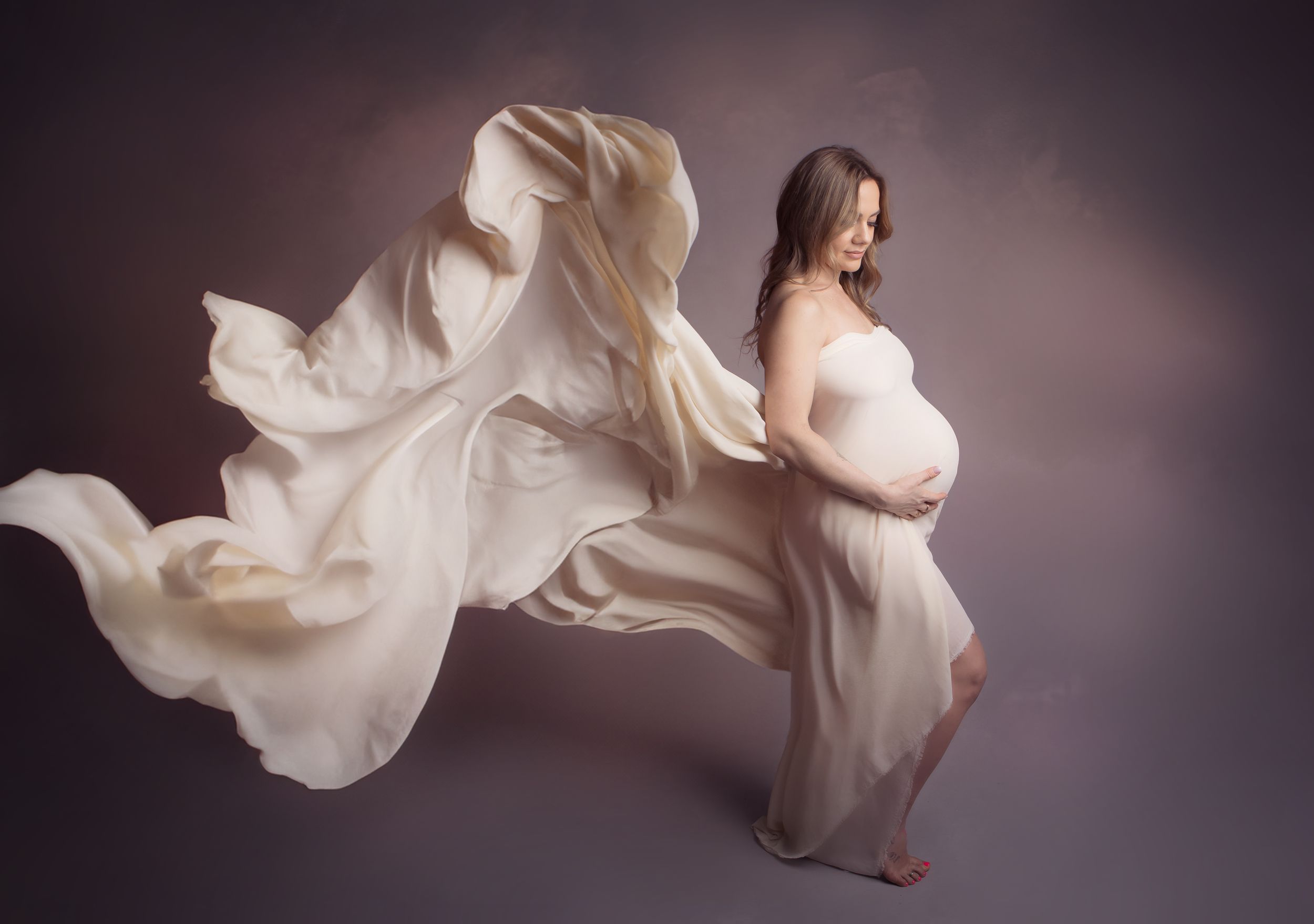 Ethereal maternity photo with flowing white fabric against moody studio backdrop creating dramatic movement and texture.