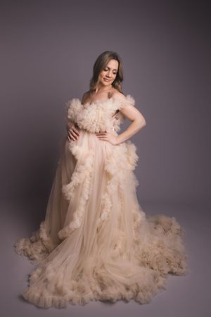 A woman poses gracefully in a romantic blush tulle gown with ruffled details and flowing train against a gray backdrop.