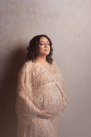 Maternity photo of woman in sequined champagne gown against neutral backdrop.
