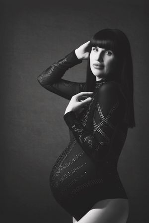 A moody black and white photoshoot shows a striking figure in black posing artistically against a dark background.
