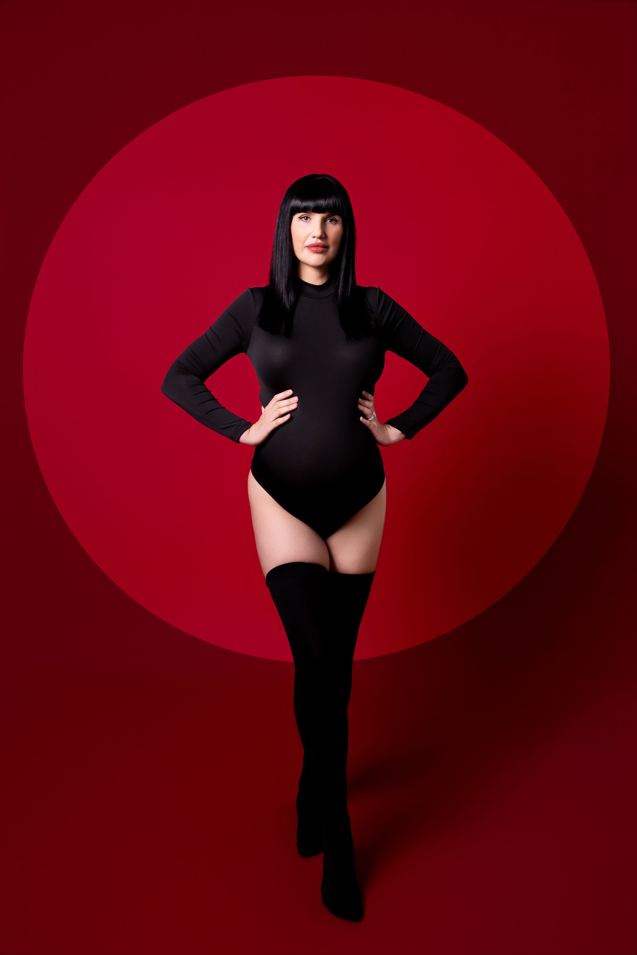 A figure in a black bodysuit poses dramatically against a deep red circular backdrop in a stylized fashion series.