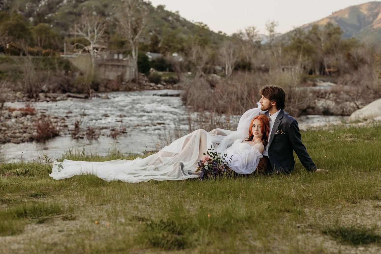 A couple sits together on grass beside a river with mountains in the background at sunset.