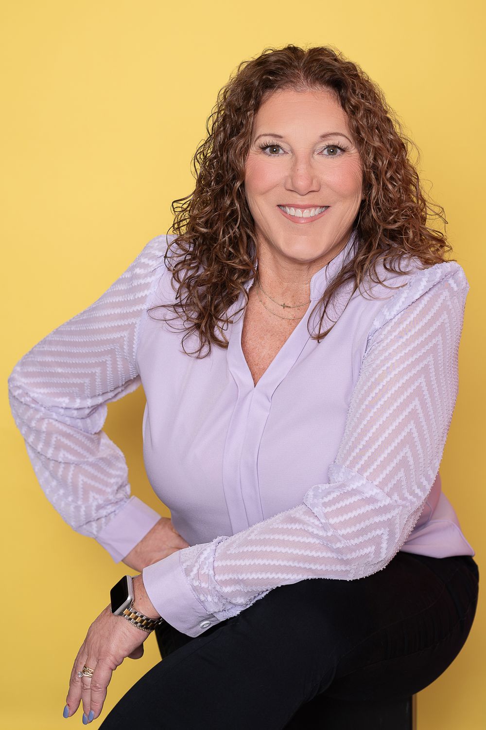 Professional headshot of someone in a lilac blouse with sheer striped sleeves against a yellow background.