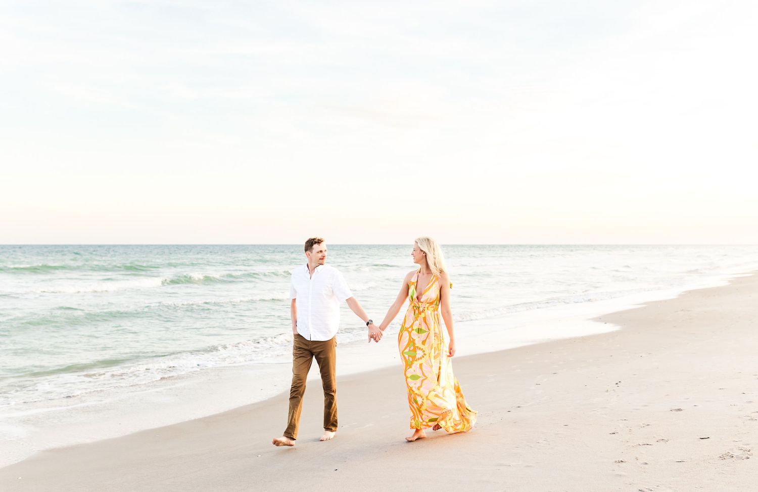 A couple in casual attire walks hand in hand along a serene beach at sunset.