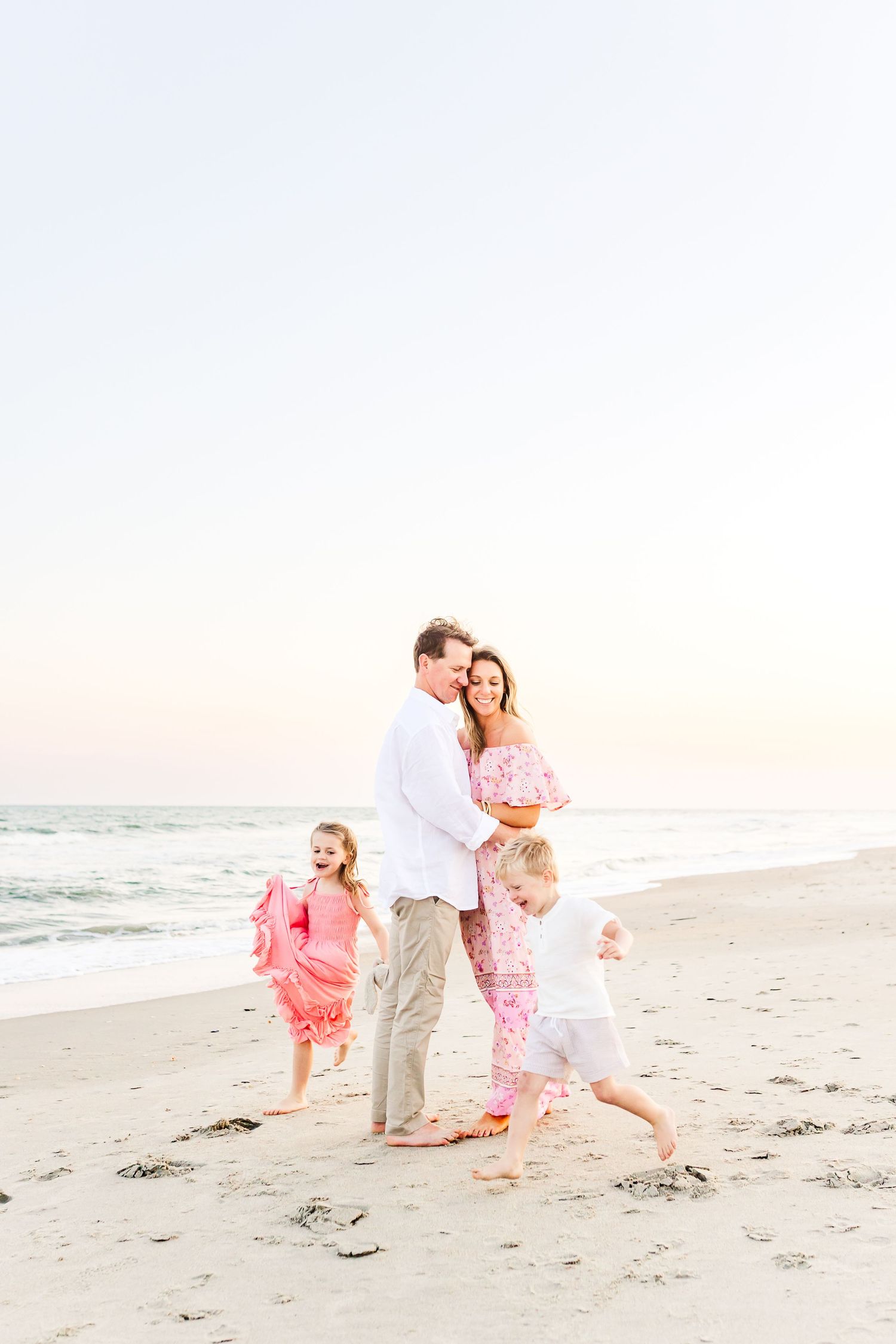 A family in coordinating pink and white outfits walks together on a sunny beach at sunset.