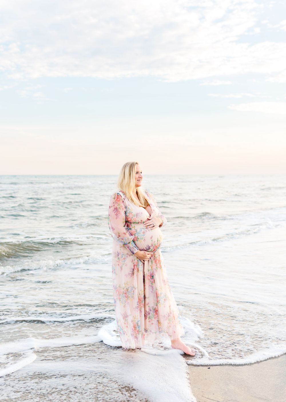 Woman in flowing pink floral dress stands gracefully at ocean's edge during sunset photoshoot on sandy beach.