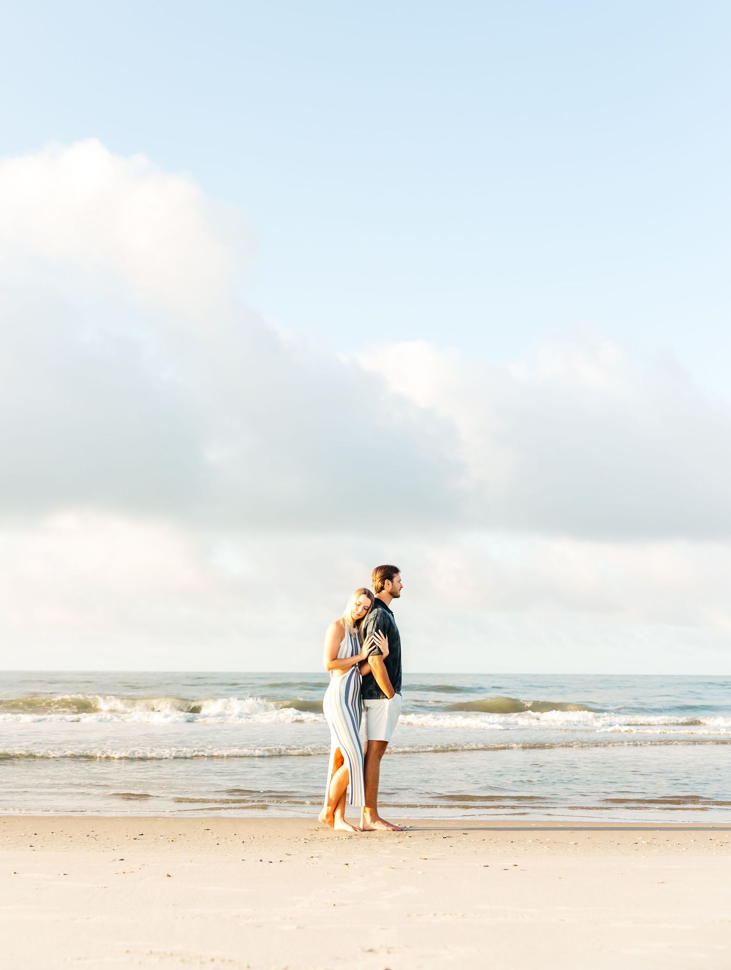 Couple embraces while standing on beach with waves rolling in. PRESLEY ROSE PHOTOGRAPHY OCEAN ISLE BEACH SUNSET BEACH BRUNSWICK BEACHES NC