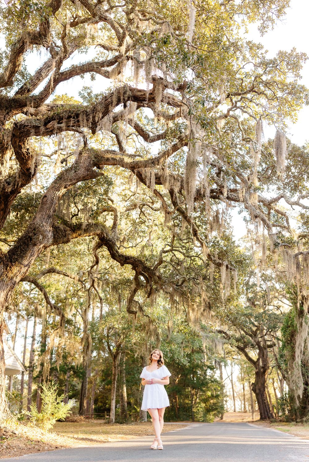 Woman in white dress walks under majestic oak trees draped with Spanish moss. PRESLEY ROSE PHOTOGRAPHY OCEAN ISLE BEACH SUNSET BEACH SUPPLY