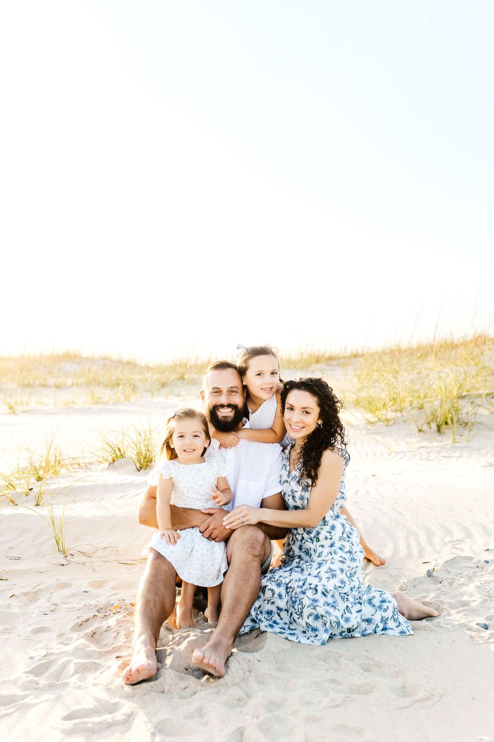 A family sits together on a sandy beach during golden hour, wearing blue and white outfits.