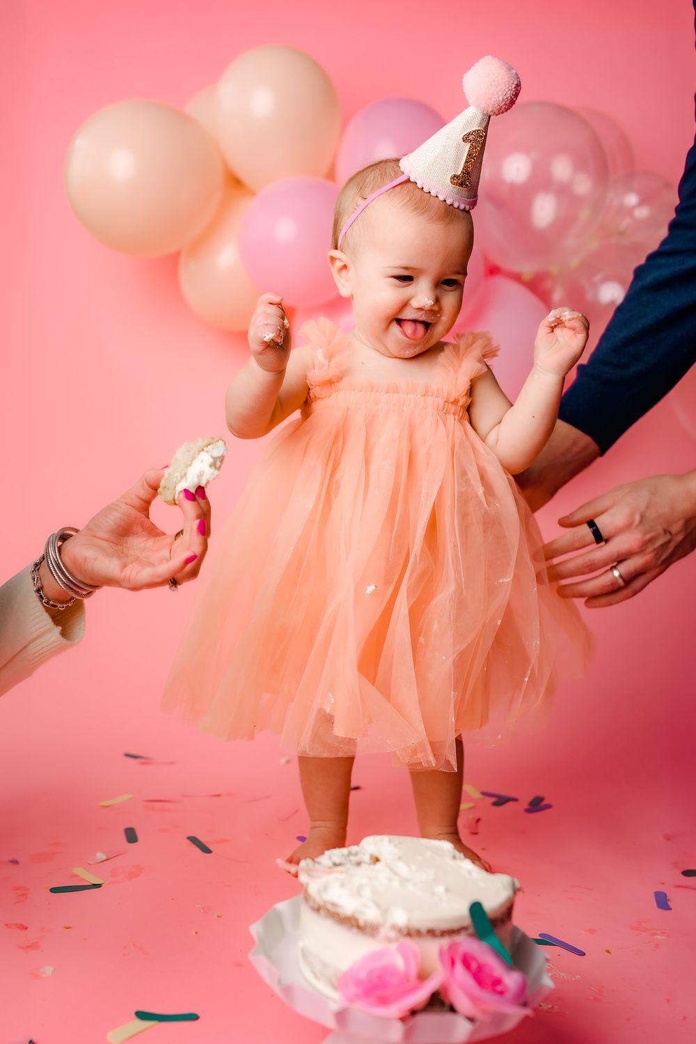 Happy baby girl in peach dress and party hat celebrates first birthday with cake and pink balloons on pink background.