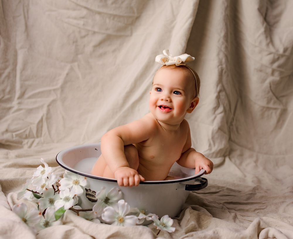Smiling baby sitting in a vintage enamel wash basin with white flowers, wearing a floral headband.