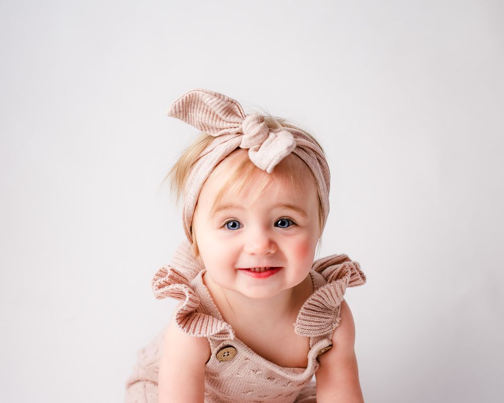 Smiling baby girl in pink knit romper and matching bow headband poses against white background.