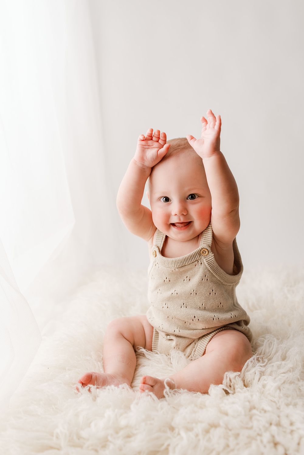 Smiling baby in beige knit romper sitting on fluffy white rug with arms raised joyfully.