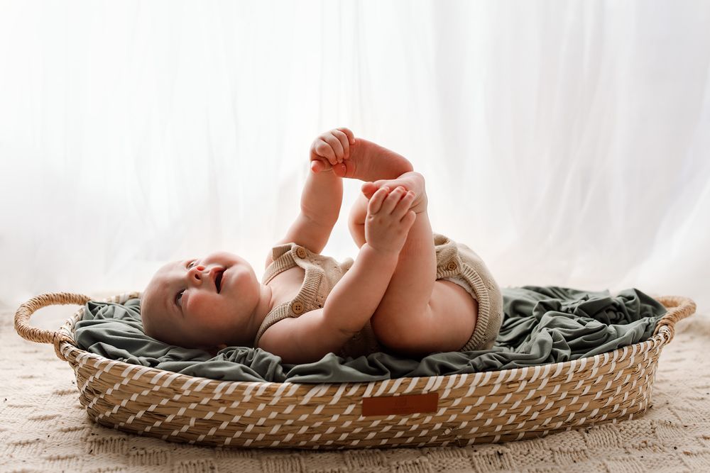A happy baby lying in a woven Moses basket on a blanket, playfully grabbing their feet against a white backdrop.