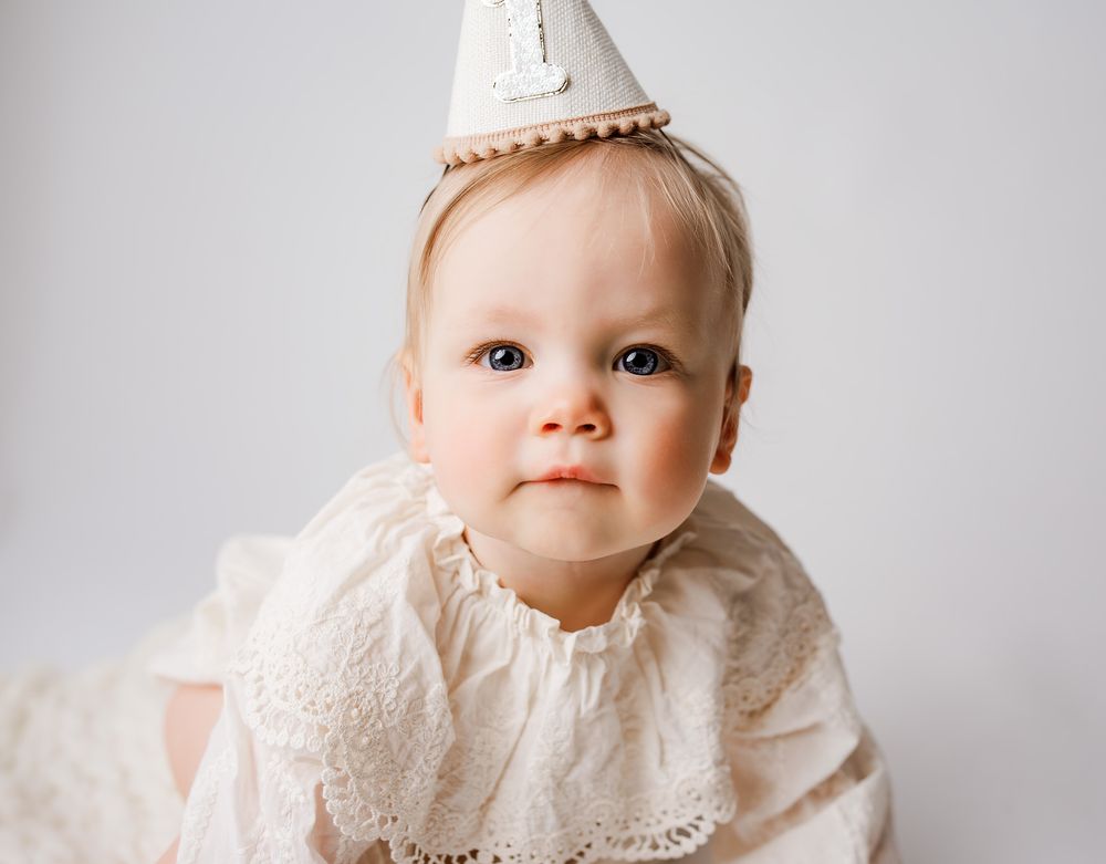 Adorable baby girl in white lace dress and birthday party hat celebrating her first birthday.