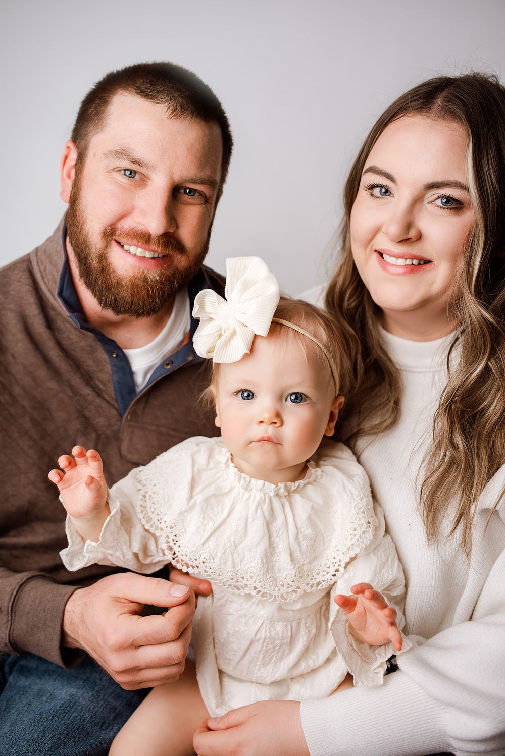 A smiling couple poses with their baby girl in a white bow and dress in a bright studio family portrait.