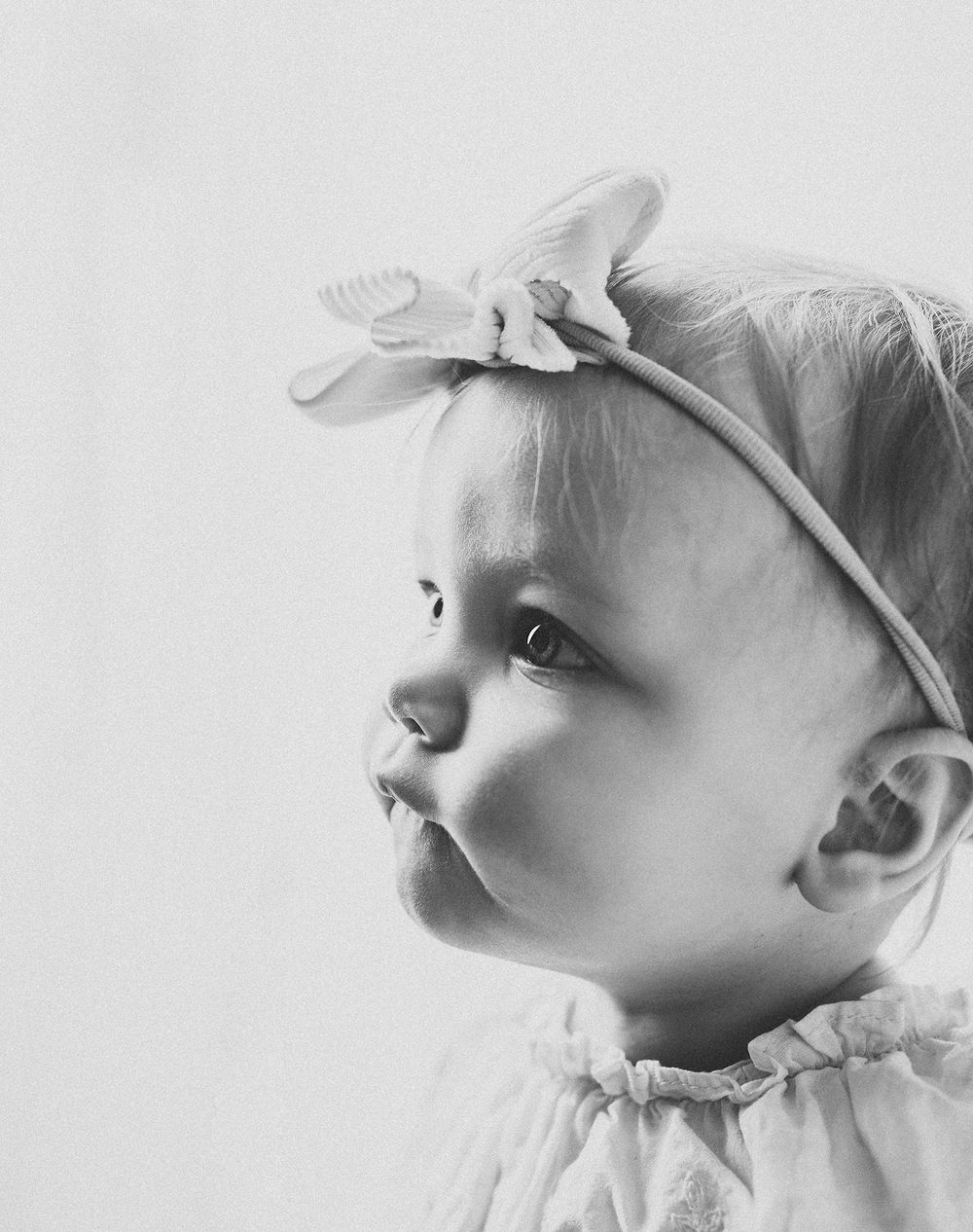 Black and white portrait of a cute baby girl wearing a floral headband, looking sideways with a curious expression.