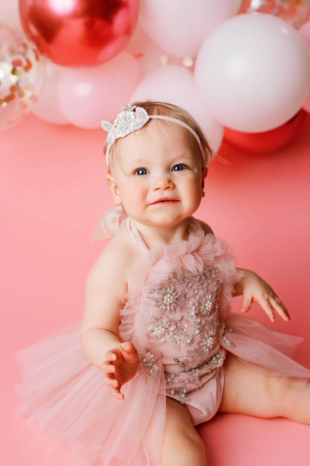 Adorable baby girl in pink tutu dress and jeweled headband sitting against pink background with balloons.
