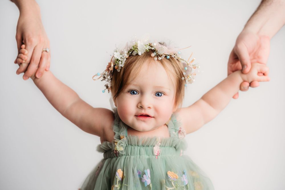 Adorable toddler girl in green floral tulle dress with flower crown held by parents hands in studio.