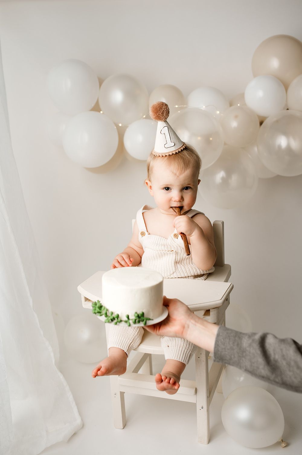 Baby in beige romper wearing birthday hat sits in white high chair with cake, surrounded by white balloons.