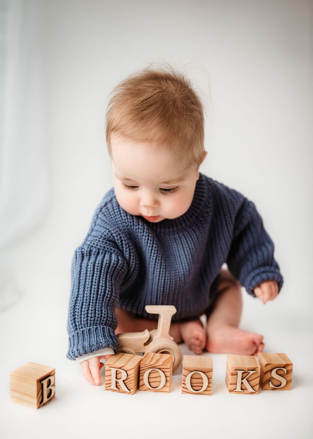 Baby in navy sweater playing with wooden letter blocks spelling BROOKS on white background.