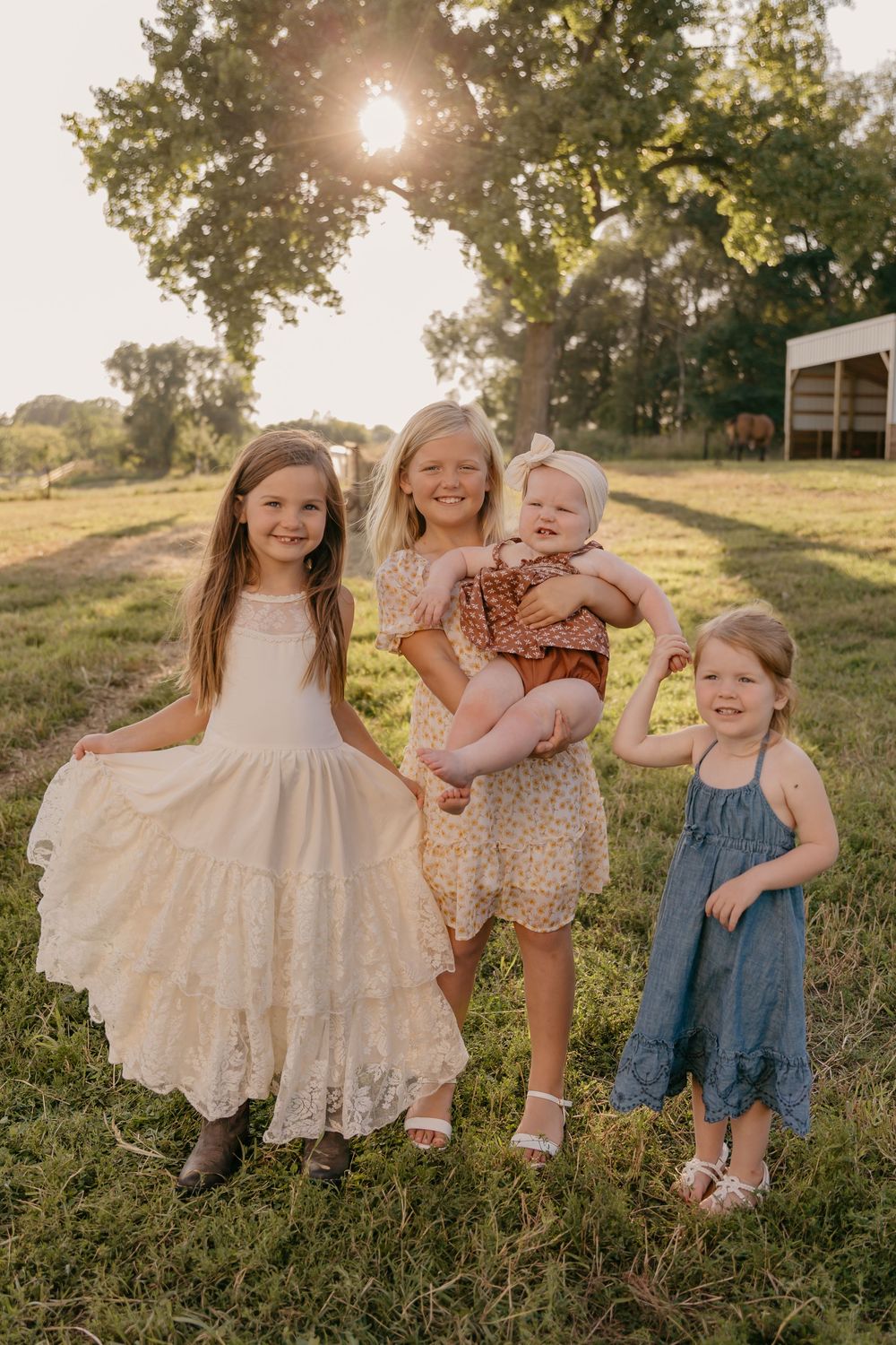 Children in summer dresses stand together in a field at sunset with sunlight peeking through trees in the background.