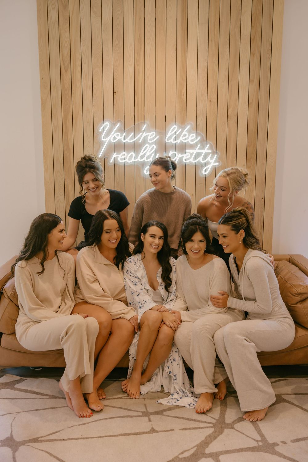 A group of women in neutral-colored loungewear gather on a couch beneath a neon sign.