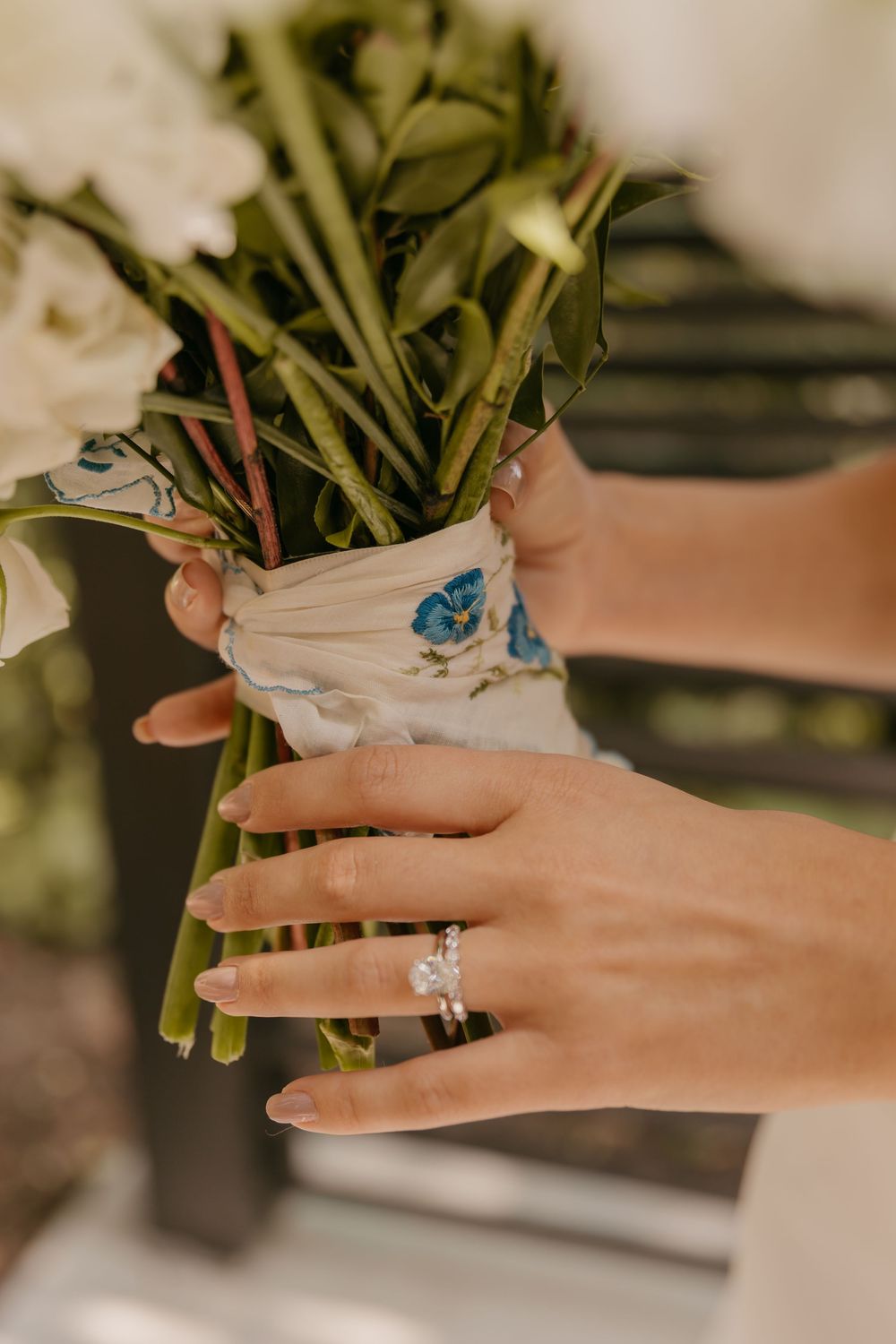 Close-up of hands holding a bridal bouquet, showcasing engagement ring and blue floral details.