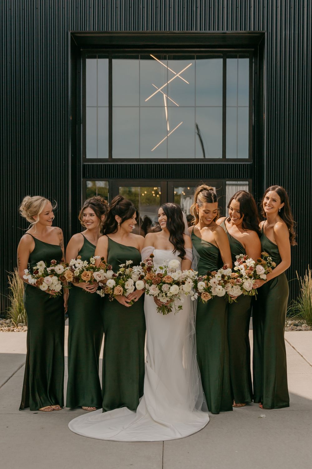 Wedding party in elegant emerald green bridesmaid dresses pose with bride in white gown holding lush floral bouquets.