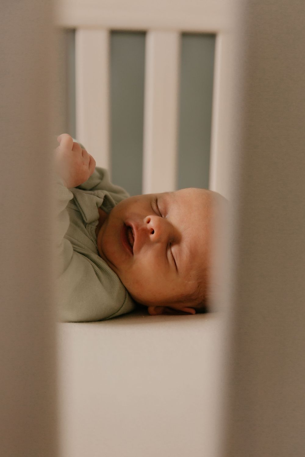 A sleeping baby in green clothing viewed through white crib slats in a softly lit room.