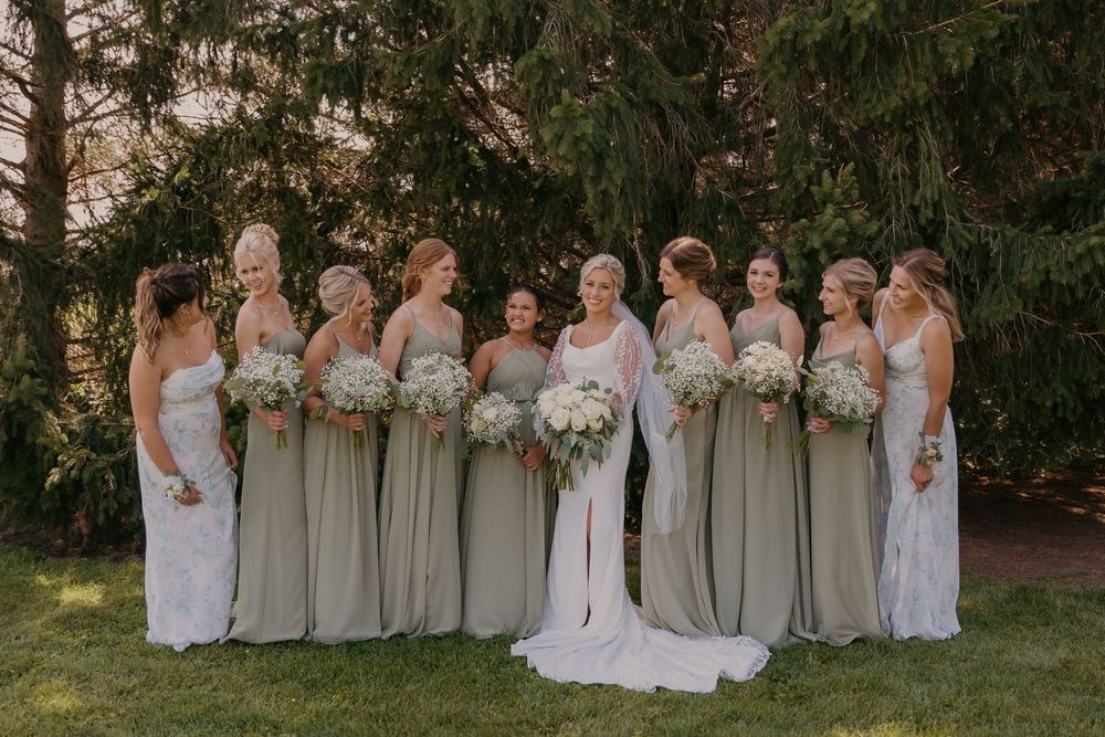 A wedding party with bridesmaids wearing sage green dresses poses outdoors against evergreen trees.