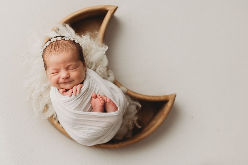 Sleeping newborn wearing white bow headband rests peacefully in black round basket with lace trim on wooden floor.