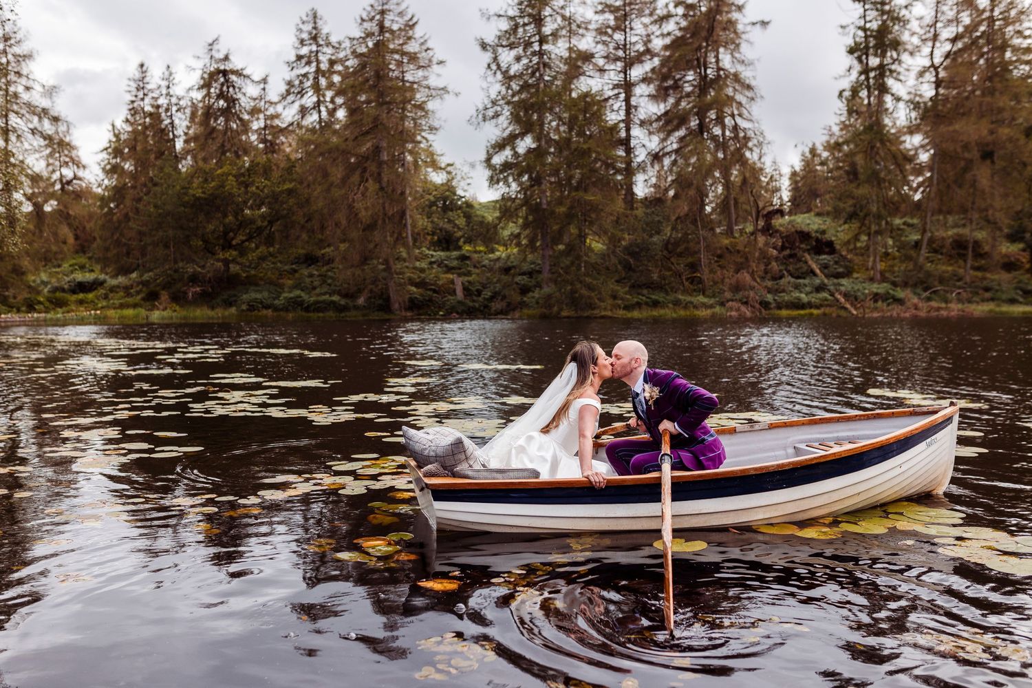 Newlyweds enjoying a romantic boat ride on a lake surrounded by pine trees.