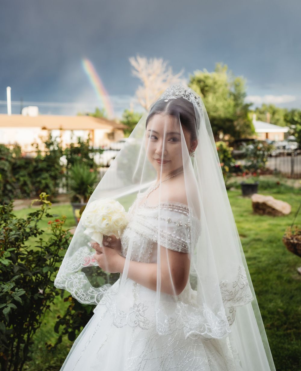 Bride covered in her veil over her face standing in front of a rainbow for solemn portrait before backyard wedding ceremony in Albuquerque.