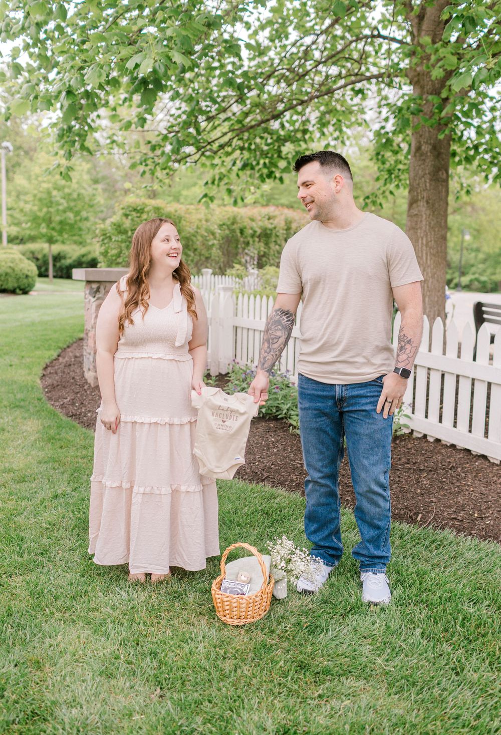 A couple smiling at each other outdoors, woman in pink maxi dress, man in beige shirt and jeans, holding baby clothes.
