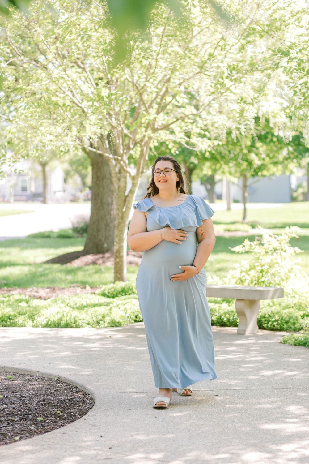 Pregnant woman in light blue maxi dress cradling baby bump while walking through a sunny park.