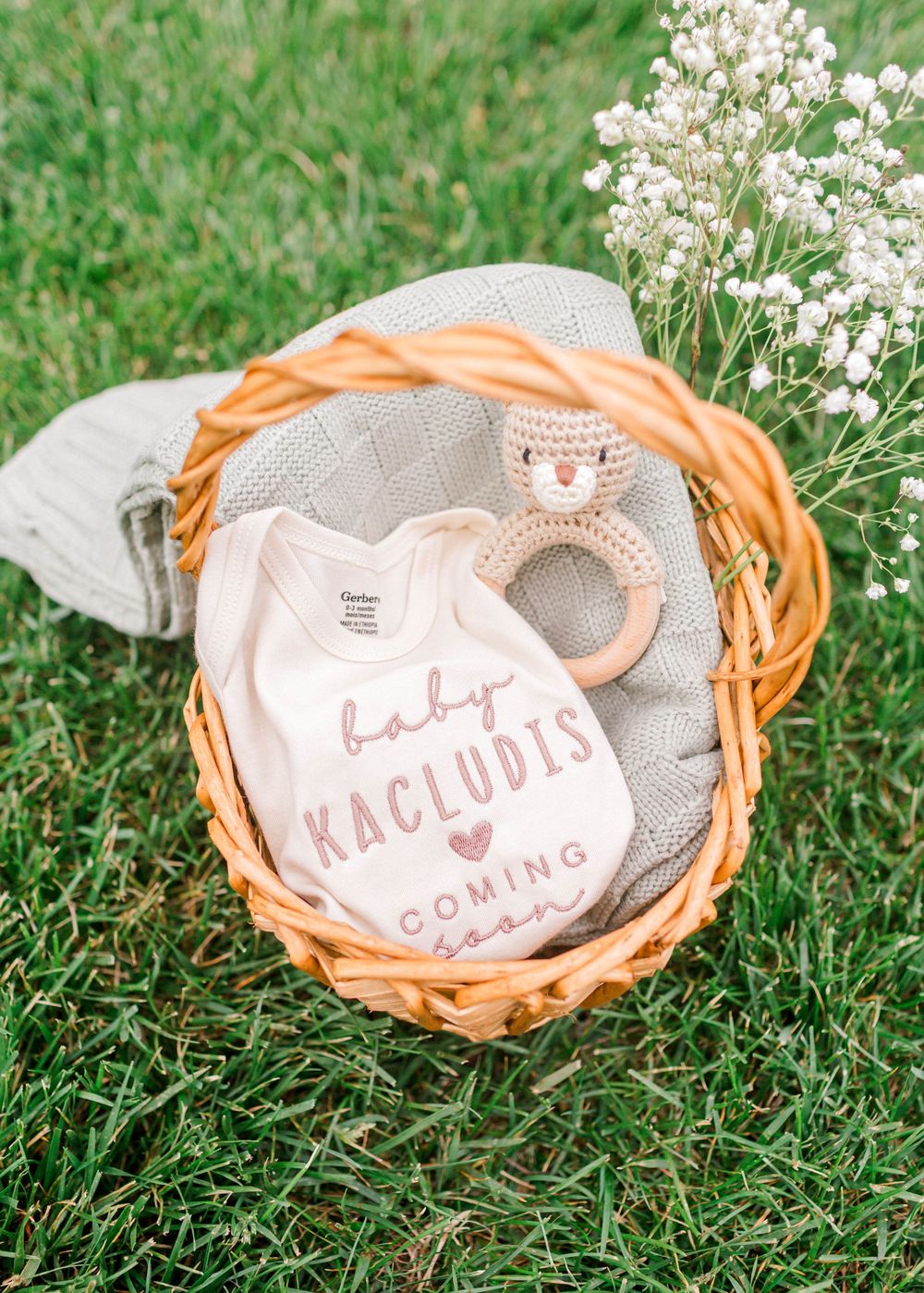 Baby announcement onesie in a wicker basket with crochet toy and baby blanket on green grass.