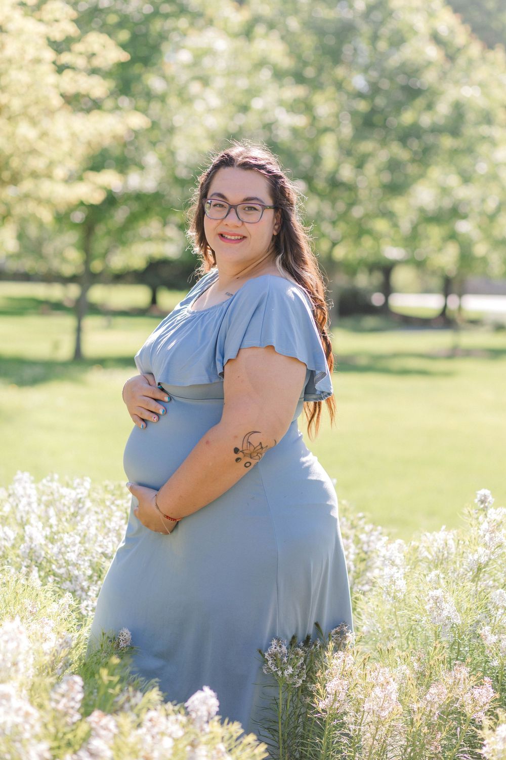 Pregnant woman in blue dress holding baby bump outdoors among wildflowers in a sunny park setting.