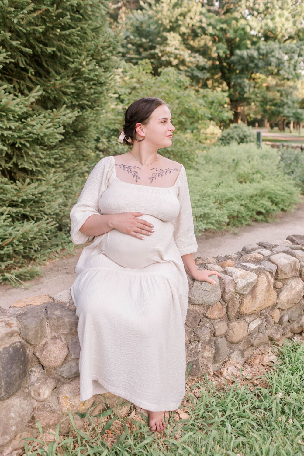 Pregnant woman in white dress sitting on stone wall in lush green park, cradling baby bump.
