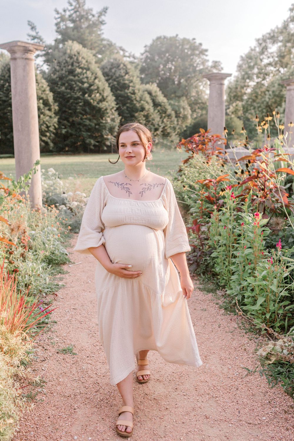 Pregnant woman in cream dress walks garden path with colorful flowers and stone columns in soft sunlight.