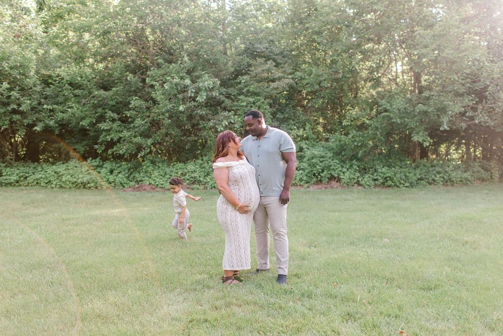 Pregnant Black woman in white dress with husband and toddler in sunny green park during maternity photo session.