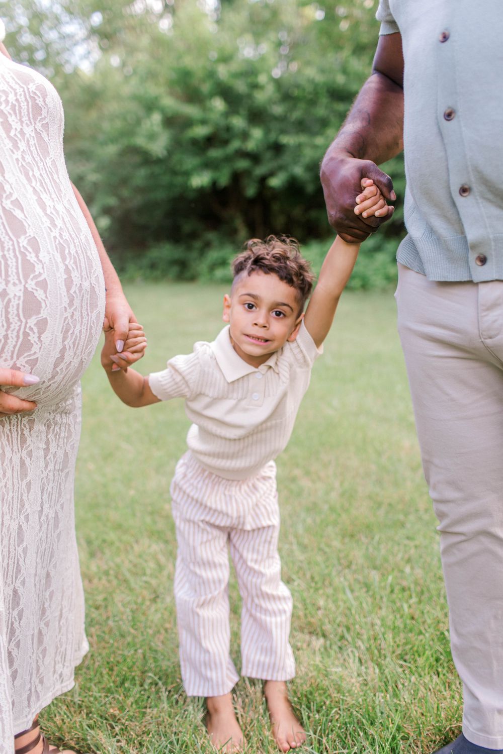 Young boy holds parents hands outdoors, standing between pregnant mother in lace dress and father in a green field.