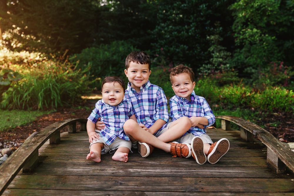 Three children in coordinating blue and white outfits sit together on a wooden bridge in a garden setting.