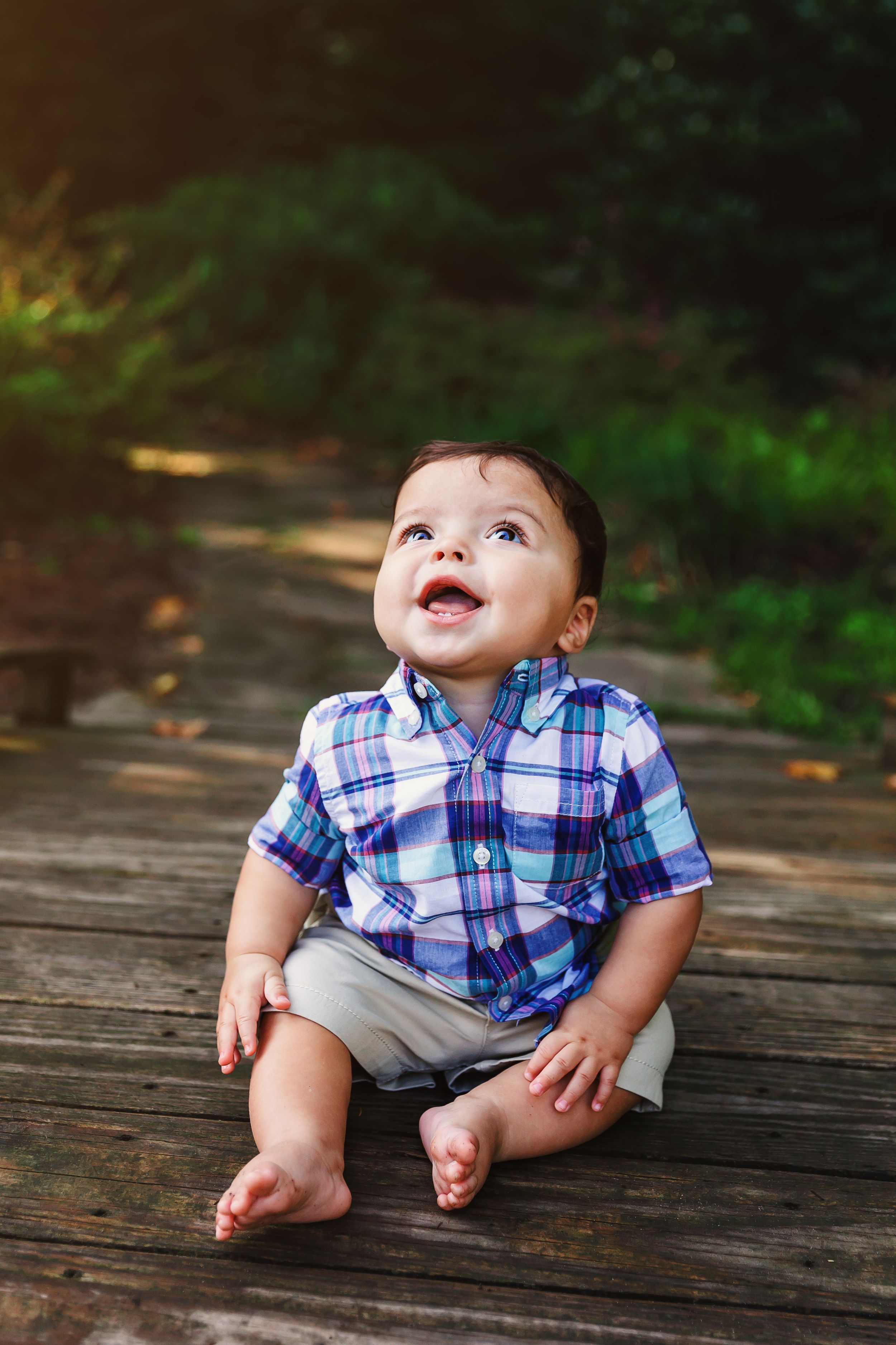 A baby wearing a blue plaid shirt and khaki shorts sits on a wooden deck with a joyful expression.
