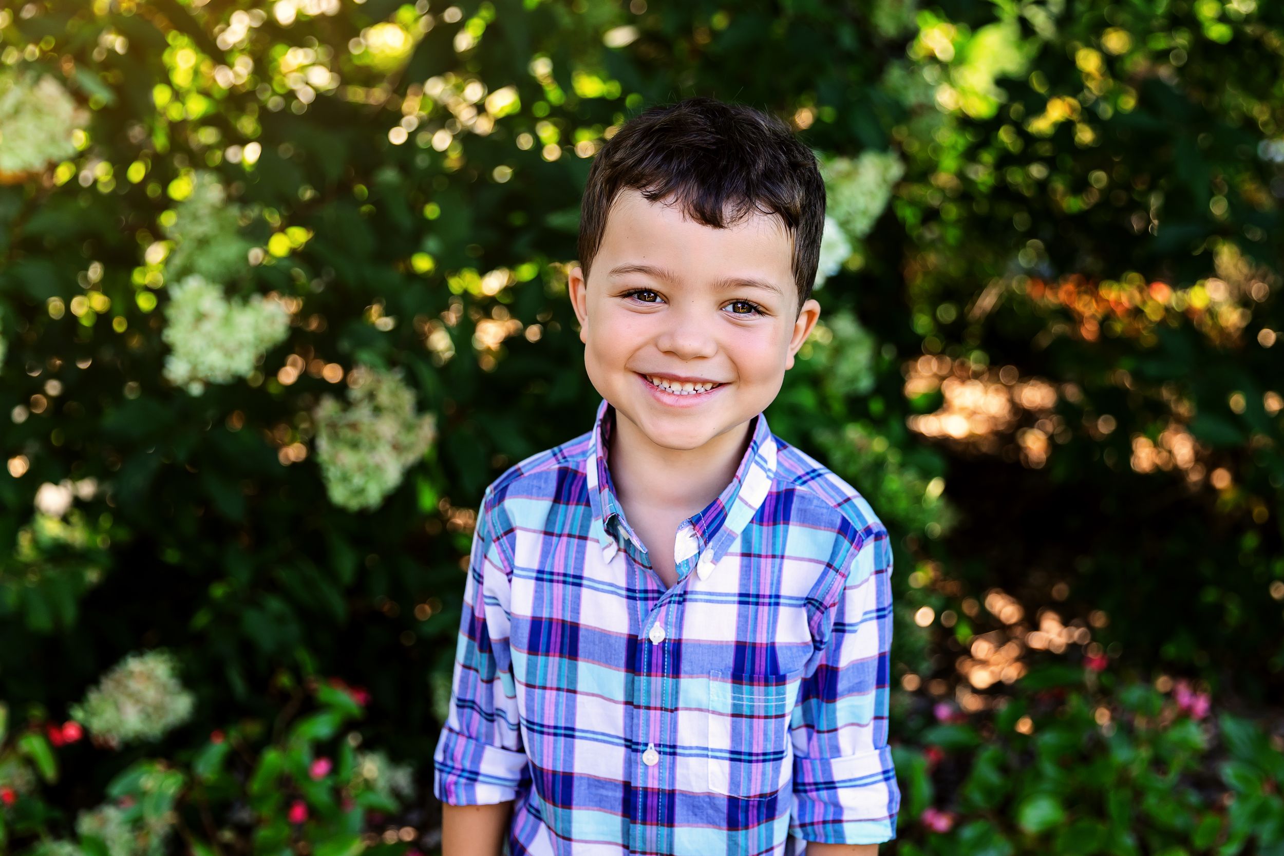 A person in a blue plaid shirt smiles outdoors against a green leafy background in natural lighting.