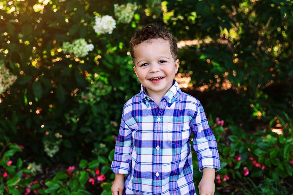 A child wearing a blue and white plaid shirt smiles while posing outdoors in a natural setting with greenery.