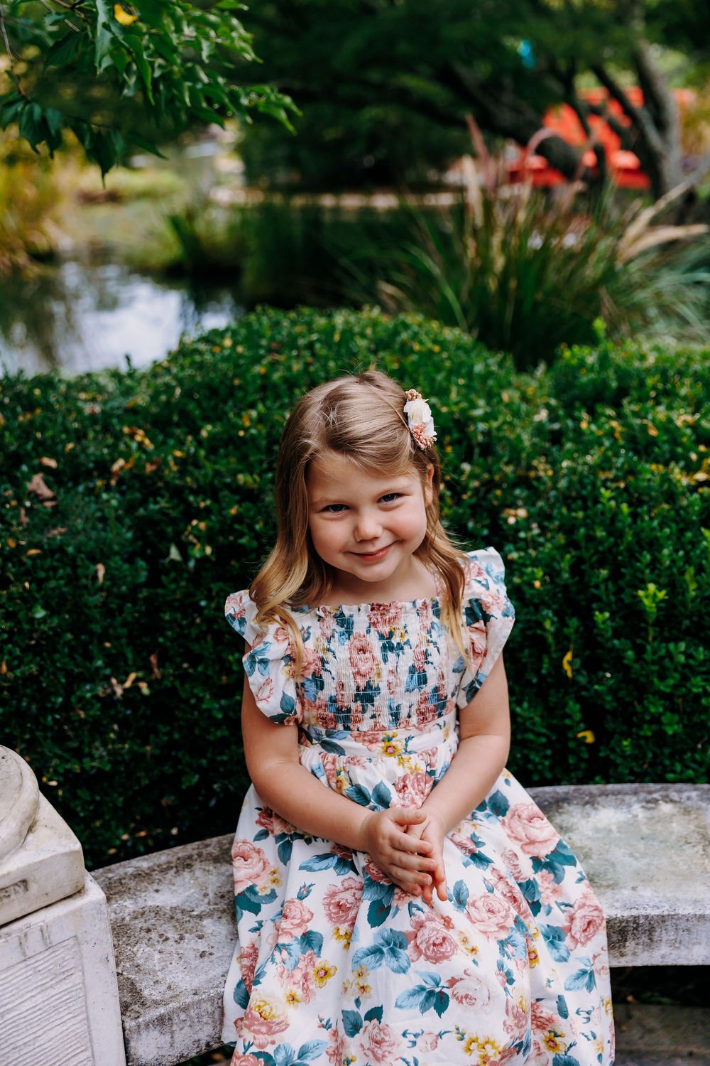 A child in a floral print dress sits on a stone bench in a garden setting with lush greenery and water in the background.